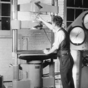 A black and white phot of a man in shirtsleeves and vest, his back to us, fiddling with a large machine.