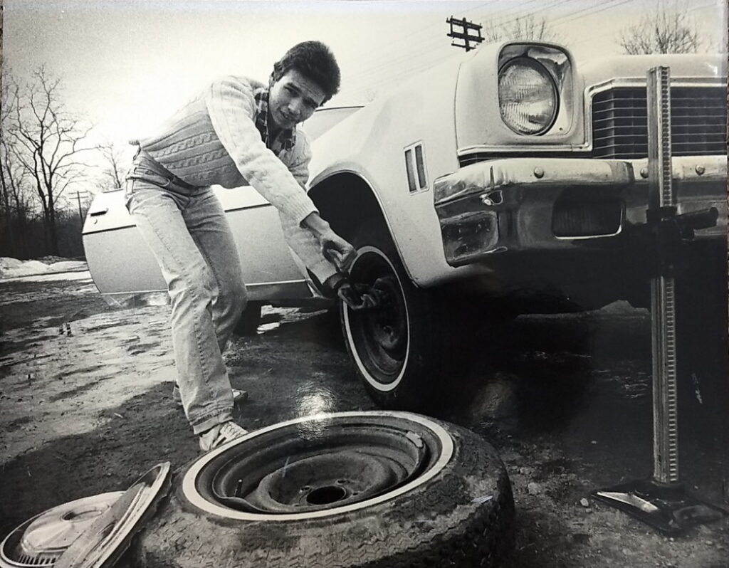 Black and white photograph of a young man changing the tire on a white car, ca. 1970s