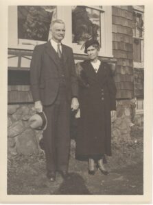 Sepia full-length photo of a man and women standing in front of a fieldstone house.